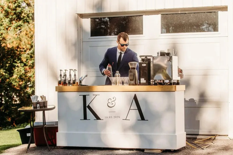 A barista in a suit making drinks at the Riverbond coffee cart, decorated with the couple’s initials, at an outdoor wedding