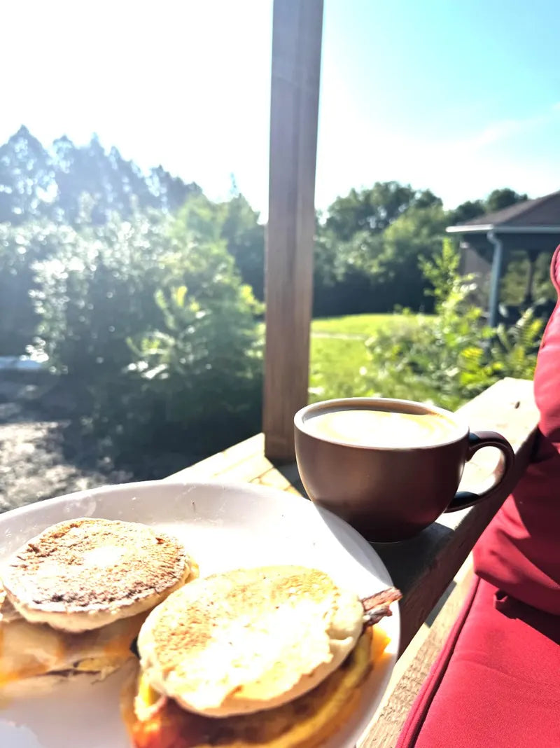 A latte in a brown cup next to breakfast sandwiches on a sunny porch railing